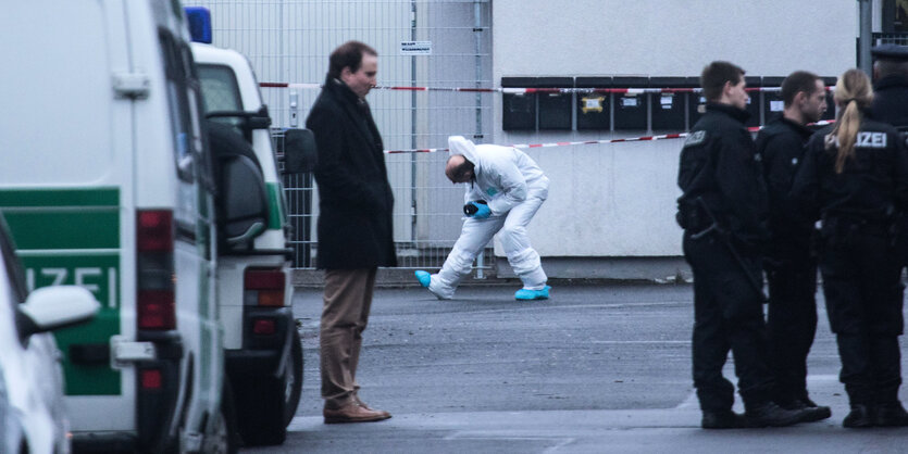 Polizisten stehen auf einem Parkplatz herum, im Hintergrund ein Mann im weißen Overall bei der Sicherung von Spuren.