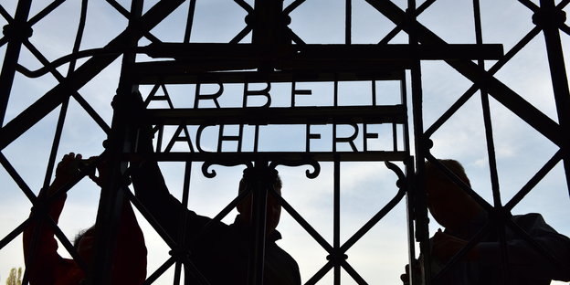 drei Arbeiter montieren „Arbeit mach Frei“ an das KZ-Tor von Dachau