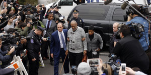Bill Cosby mit Gehstock, gestützt von zwei Helfer_innen, umringt von einer Menschenmenge