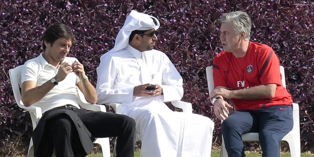 Nasser al-Khelaifi sitzt zwischen zwei Männer auf grünem Rasen