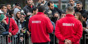 zwei Security-Mitarbeiter stehen vor einer Absperrung vor vielen Flüchtlingen