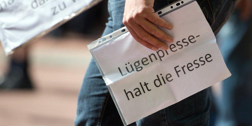 Eine Person hält einen Zettel, der sich in einer Schutzhülle befindet. Auf ihm steht: „Lügenpresse halt die Fresse".