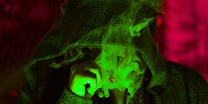 grün beleuchtete Person mit ins Gesicht gezogener Kapuze und Rauchschwaden vor dem Gesicht