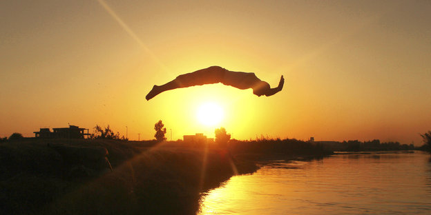 Ein Mann springt in einen langgestreckten Sprung ins Wasser, hinter ihm steht tief die Sonne.