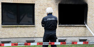 Polizist steht hinter rot-weißen Absperrband vor einer Hausfassade mit ausgebrannter Fensterhöhle.