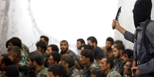 Ein IS-Kämpfer hält vor sitzenden Männern ein Messer in Höhe