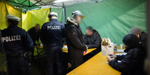 Polizisten kontrollieren Verdächtige an Tischen in einem grünen Zelt