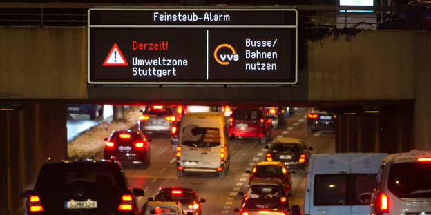 Autos von hinten auf einer Autobahn, darüber digitales Schild, auf dem eine Feinstaub-Warnung zu lesen ist und die Aufforderung, Bus und Bahn zu nutzen
