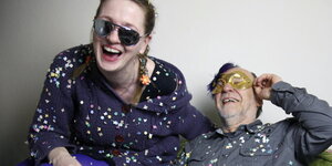 junge Frau mit Sonnenbrille und ein älterer Mann mit Maske sitzen auf einem Sofa und lachen. Sie haben Konfetti auf ihrer Kleidung