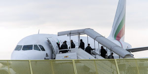 Hinter einem Zaun gehen Menschen über eine Gangway in ein Flugzeug