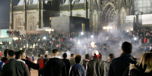 Menschen stehen in einer Ansammlung vor dem Kölner Dom, Rauchschwaden hängen in der Luft