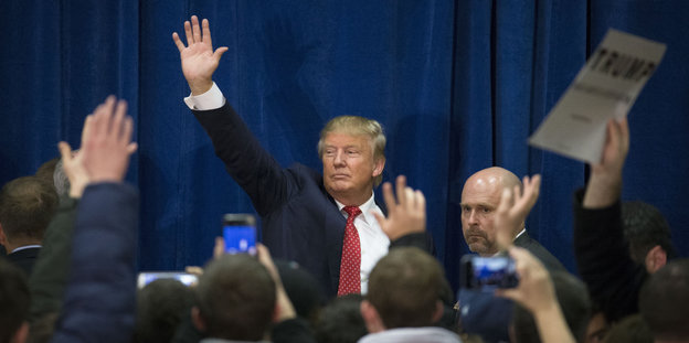 Mann im Anzug, es ist Donald Trump, hebt seine Hand, vor ihm lauter Menschen, teilweise auch mit in die Höhe gestreckten Armen