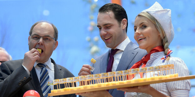 Ein Mann im grauen Anzug beißt in ein Stück Käse, neben ihm stehen ein Mann in einem hellgrauen Anzug und eine Frau in niederländischer Tracht, die ein Brett mit Käsewürfeln hält