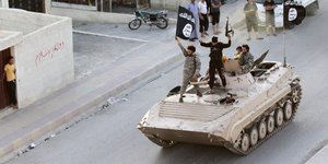 IS-Kämpfer stehen auf einem Panzer, der eine Straße entlang fährt. Einer von ihnen schwenkt eine IS-Fahne.