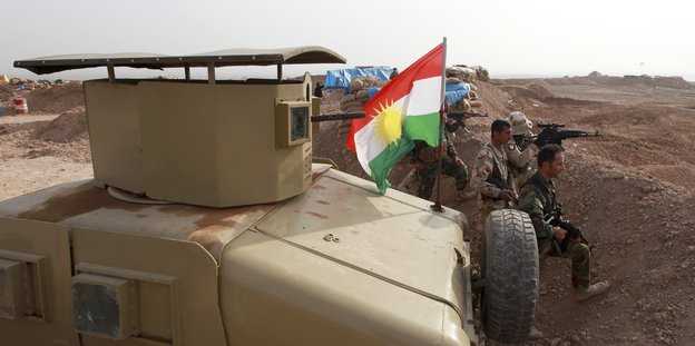 Ein Panzer mit einer kurdischen Flagge, davor in einem Schützengraben Soldaten