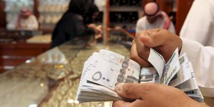 Im Vordergrund halten Hände ein Geldscheinbündel, im Hintergrund ein Geschäft in Saudi-Arabien