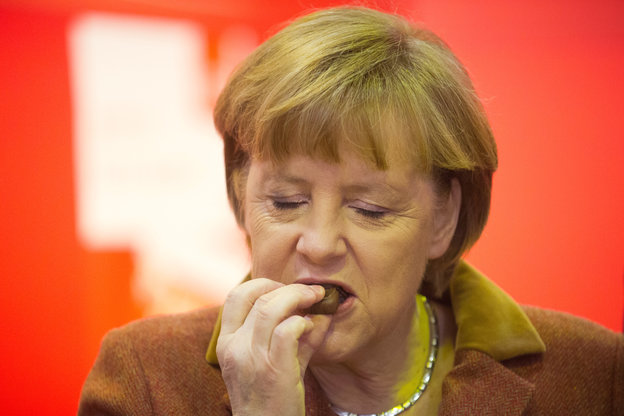 Bundeskanzlerin Angela Merkel isst genüsslich eine Schokopraline vor rot-weißem Hintergrund
