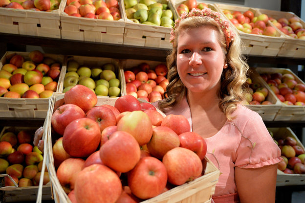 Blonde junge Frau hält einen Korb mit roten Äpfeln im Arm. Im Hintergrund ein Obststand.