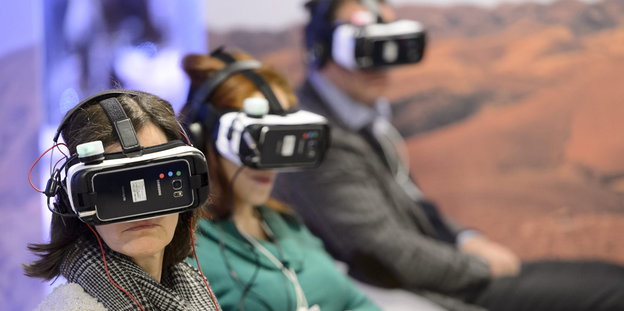Drei Personen sitzen auf einer Bank und haben eine Virtual-Reality-Brille auf.