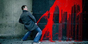 Ein Mann steht in Pinkelpose vor einer mit rotem Graffiti besprühten Wand