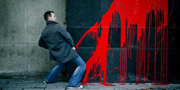 Ein Mann steht in Pinkelpose vor einer mit rotem Graffiti besprühten Wand