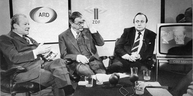 Brandt, Kohl und Genscher in einem Fernsehstudio, auf einem Bildschirm ist Strauß zugeschaltet