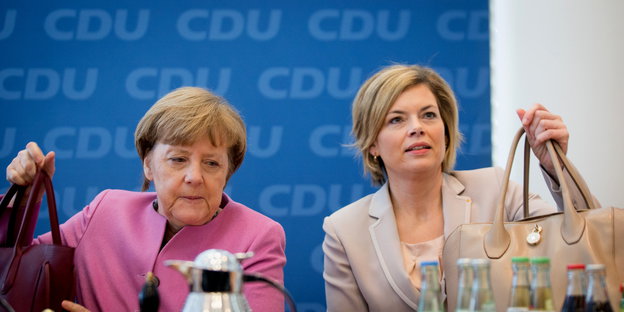 Angela Merkel und Julia Klöckner halten jeweils eine Handtasche in der Hand, im Hintergrund eine blaue Wand mit CDU-Schriftzügen