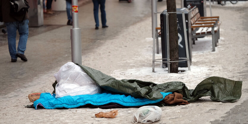 Auf einem Gehweg liegt ein Schlafsack im Schnee, dahinter eine Parkbank, Menschen laufen vorüber