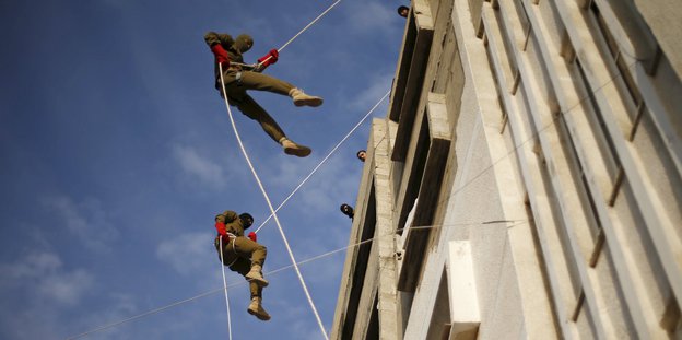 Zwei Menschen in tarnfarbener Kleidung und roten Handschuhe seilen sich von einem Gebäude ab.