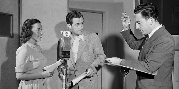 Zwei Männer und eine Frau stehen um ein Mikrofon herum – Schwarzweiß-Aufnahme