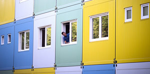 Bunte Wohncontainer, aus einem Fenster lehnt sich ein Mann und prüft den Fensterrahmen.