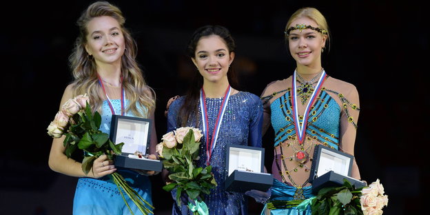 Drei Frauen stehen lächelnd nebeneinander. Sie halten Medaillen und rosafarbene Rosen in den Händen