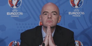 Gianni Infantino mit aneinandergelegten Händen unterm Kinn vor blauem Hintergrund mit dem Logo der EM in Frankreich