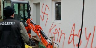 Eine Hauswand mit einem Hakekreuz und dem Spruch "Kein Asyl", davor steht ein Polizist