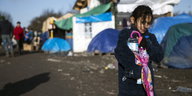 Ein Mädchen trägt einen Rehenschirm und eine Flasche Wasser durch ein Zeltlager bei Dünkirchen.