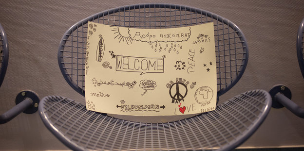 Auf einem Sitz aus Stahlgitter liegt ein handgemaltes Schild mit den Worten „Welcome“, „Peace“, daneben sind Sonne und Wolken gemalt