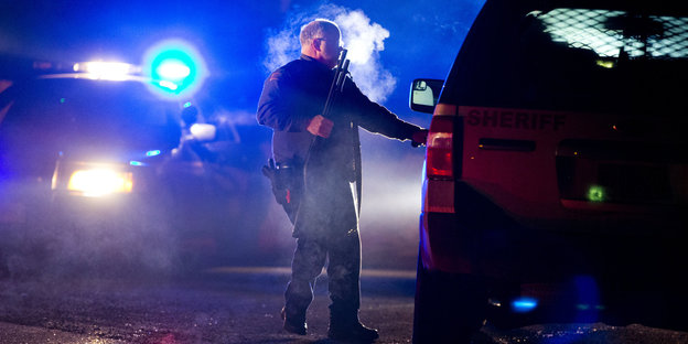 Ein amerikanischer Polizist trägt eine Waffe in ein Auto, es ist dunkel, hinter ihm Blaulicht.