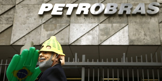 Vor dem Firmenschild „Petrobras“ steht ein Demonstrant mit einer Maske auf dem Gesicht, einem gelben Hut und einer grünen Plastikhand