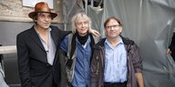 Drei Männer stehen nebeneinander: einer mit braunem Hut, einer mit Schal, einer mit hochgekrempelter Lederjacke