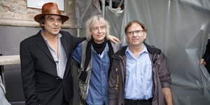 Drei Männer stehen nebeneinander: einer mit braunem Hut, einer mit Schal, einer mit hochgekrempelter Lederjacke