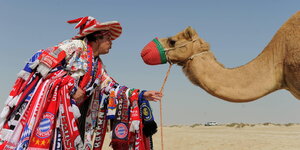 Ein Mann mit vielen Fanschals steht vor einem Kamel.