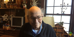 Ein alter Mann mit Brille und Galze steht lächelnd vor einem Fenster, daneben ein Computer