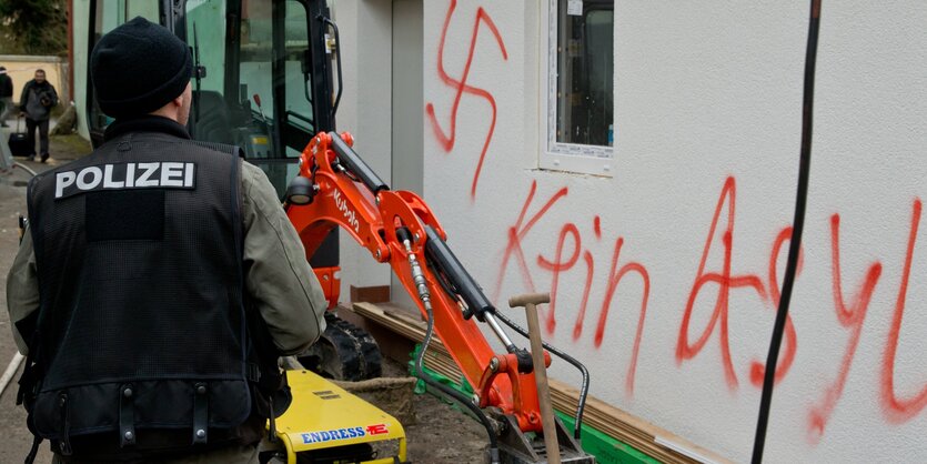 Polizist vor Fassade mit Hakenkreuzschmiererei