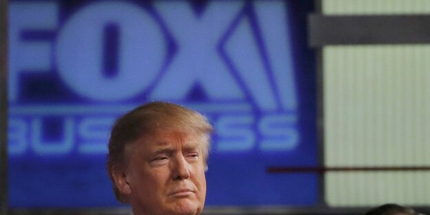 Donald Trump, ein Mann mit blonden Haaren und im Anzug, steht vor einem Schild mit der Aufschrift "Fox"