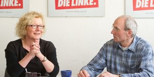 Eine Frau und ein Mann sitzen am Tisch, dahinter sind Poster der Partei Die Linke aufgehängt