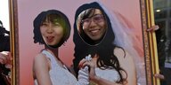 In ein Foto von zwei Frauen in Brautkleid sind zwei Löcher an Stelle der Köpfe geschnitten, durch die wiederum echte Frauen hindurchschauen und Grimassen schneiden