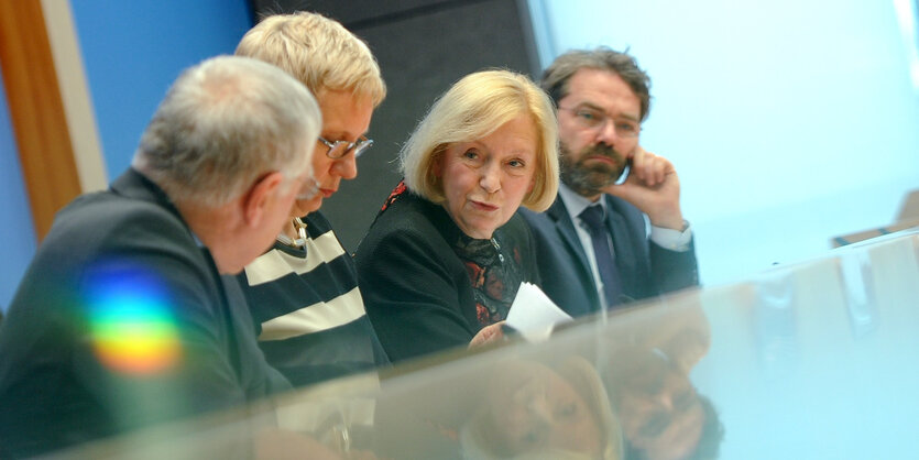 Zwei Männer und zwei Frauen sitzen an einem Glastisch, ihre Köpfe spiegeln sich in der Tischplatte