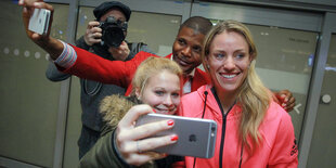 Eine strahlend lächelnde Frau mit blonden Haaren und pinker Sportjacke - es ist Angelique Kerber - vor ihr eine Frau mit Handy, hinter ihr ein Mann mit Handy, beide versuchen sich mit Kerber zu fotografieren