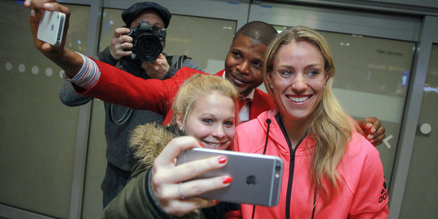Eine strahlend lächelnde Frau mit blonden Haaren und pinker Sportjacke - es ist Angelique Kerber - vor ihr eine Frau mit Handy, hinter ihr ein Mann mit Handy, beide versuchen sich mit Kerber zu fotografieren