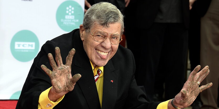 Ein Mann zeigt seine mit Zement bedeckten Hände und streckt die Zunge heraus. Es ist US-Komiker Jerry Lewis